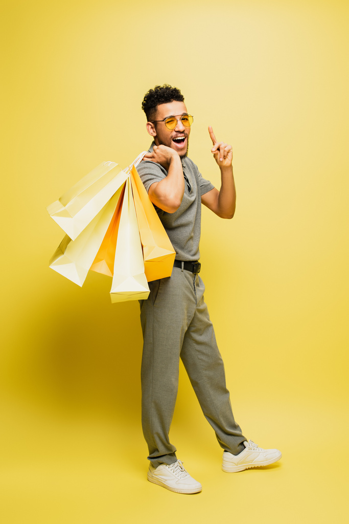in voller Länge ein fröhlicher afrikanisch-amerikanischer Mann mit Sonnenbrille und grauem Tennishemd, der Einkaufstaschen hält und auf gelb zeigt  - Foto, Bild