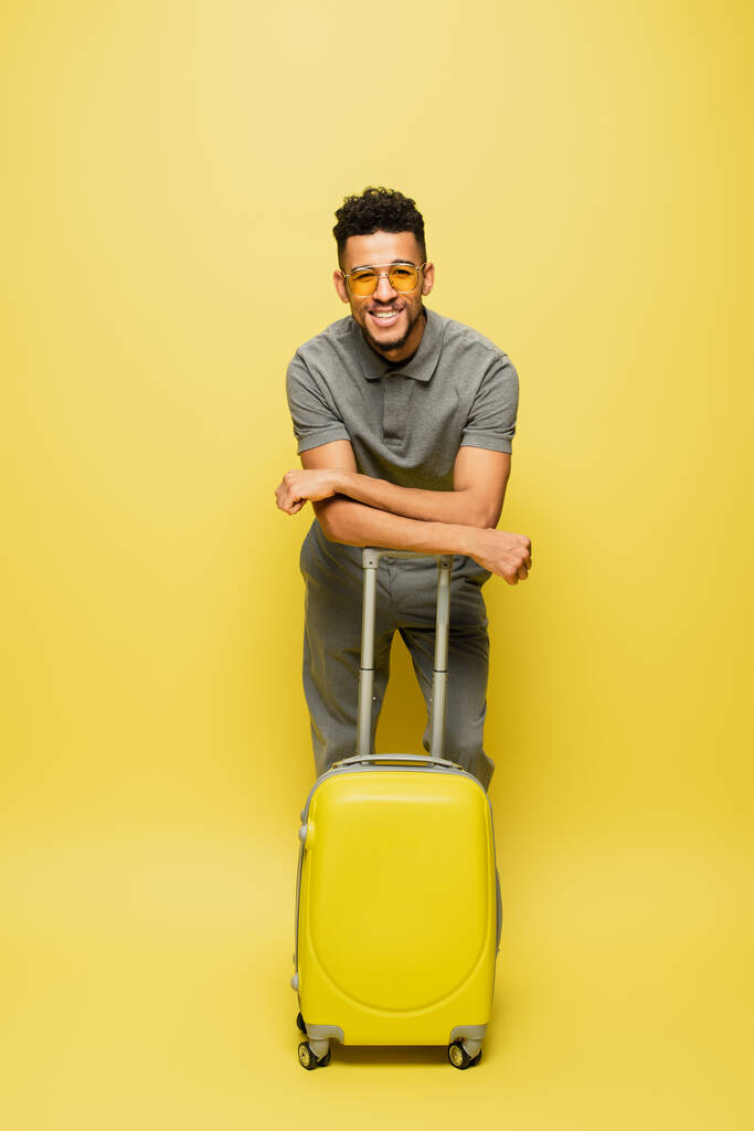 volledige lengte van gelukkig Afrikaans amerikaanse man in zonnebril en grijs tennis shirt met bagage op geel - Foto, afbeelding