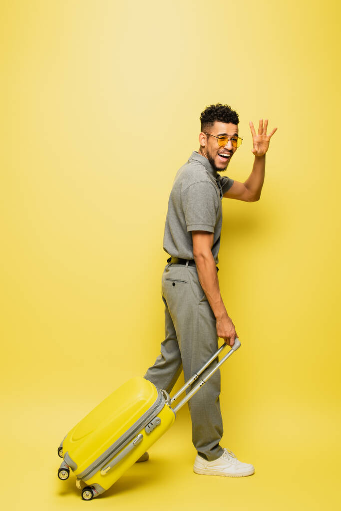 longitud completa de hombre afroamericano feliz en gafas de sol y camisa de tenis gris caminando con el equipaje y agitando la mano en amarillo - Foto, imagen