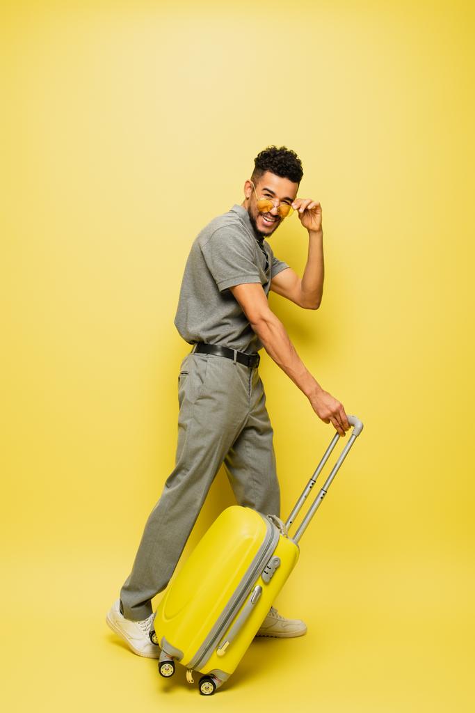 полная длина веселый молодой африканский американец регулируя солнцезащитные очки и стоя с багажом на желтом - Фото, изображение