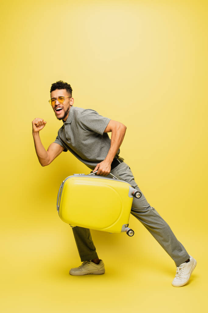 volledige lengte van opgewonden Afrikaan amerikaanse man in zonnebril en grijs tennis shirt met bagage op geel - Foto, afbeelding