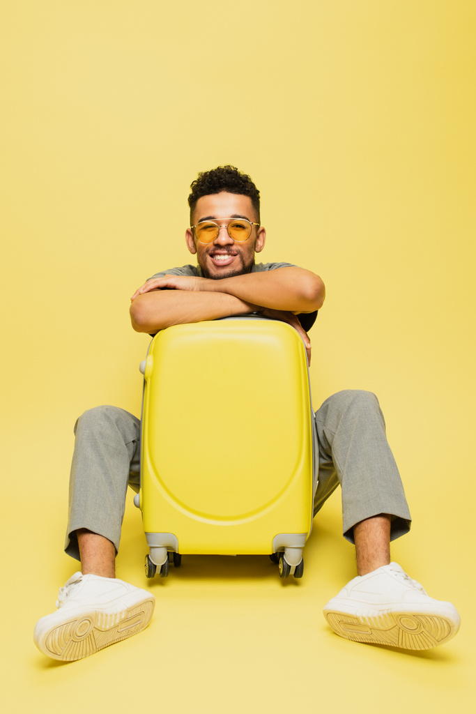 comprimento total de homem americano africano alegre em óculos de sol sentado com bagagem em amarelo - Foto, Imagem