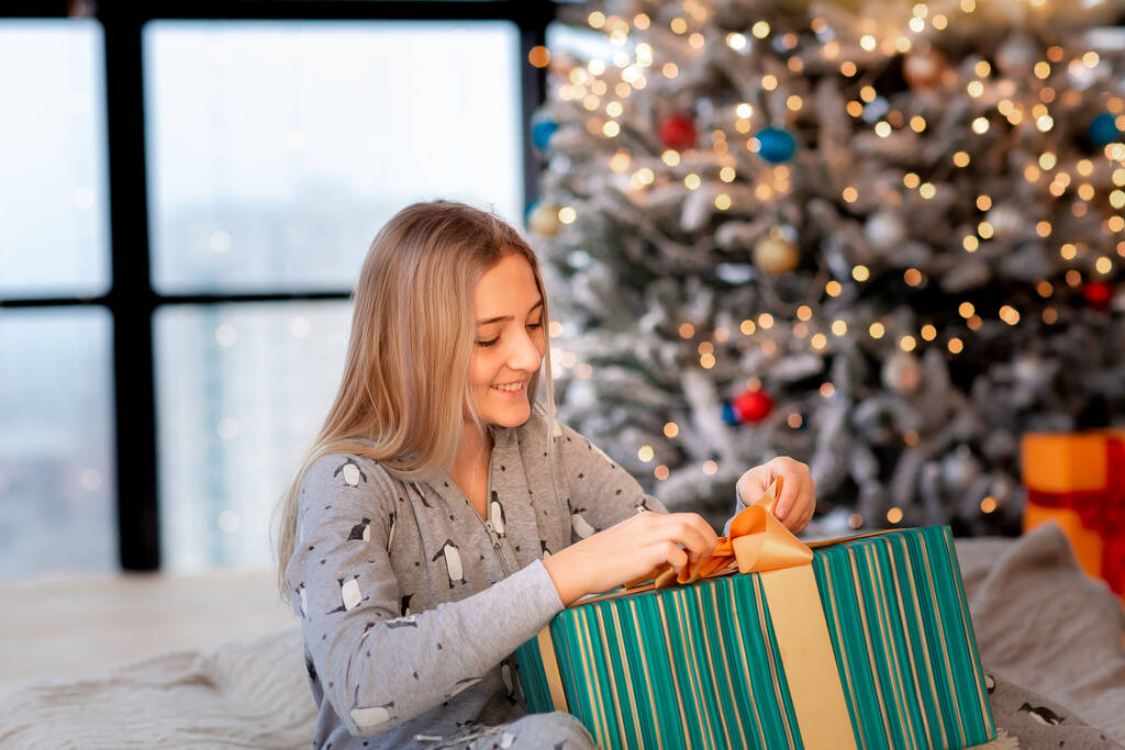 Wesoły cute teen girl otwierając prezenty. Dzieciak w piżamie bawiący się rano przy drzewie. Wesołych Świąt i Happy Holidays koncepcja - Zdjęcie, obraz