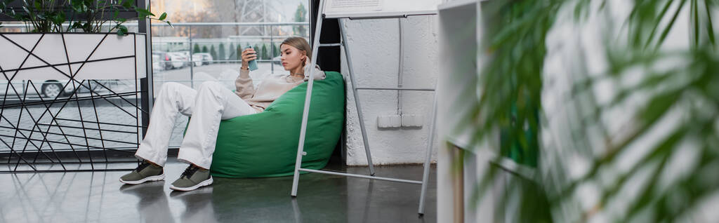 blonde manager sitting in bean bag chair and using smartphone, banner - Photo, Image
