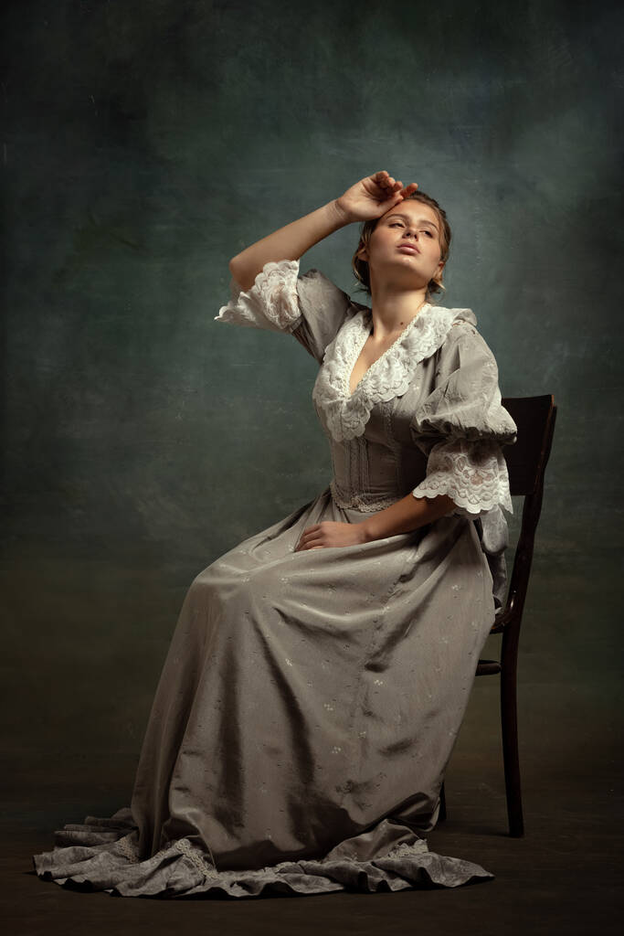 Vintage πορτρέτο του νεαρού όμορφο κορίτσι σε γκρι φόρεμα μεσαιωνικού στυλ που απομονώνονται σε σκούρο φόντο. Σύγκριση της έννοιας των εποχών, στυλ ψύλλων. - Φωτογραφία, εικόνα