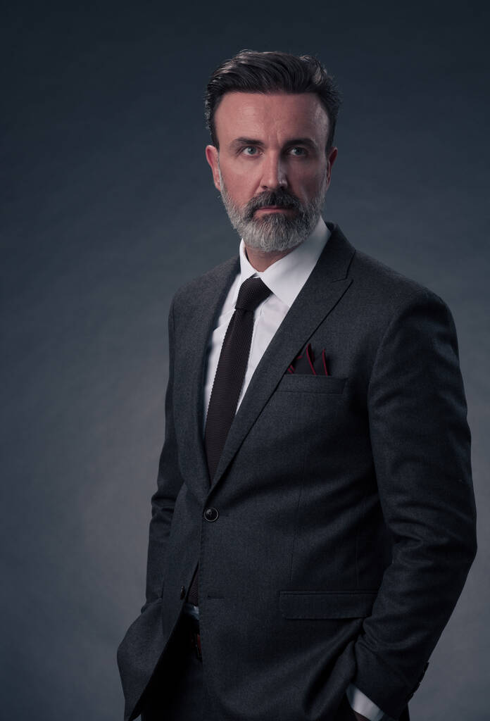Portret odnoszącego sukcesy stylowego, eleganckiego starszego biznesmena z szarą brodą i luźnymi ubraniami biznesowymi, ufnego w studio fotograficznym, odizolowanego na ciemnym tle, gestykulującego dłońmi. Wysokiej jakości zdjęcie - Zdjęcie, obraz