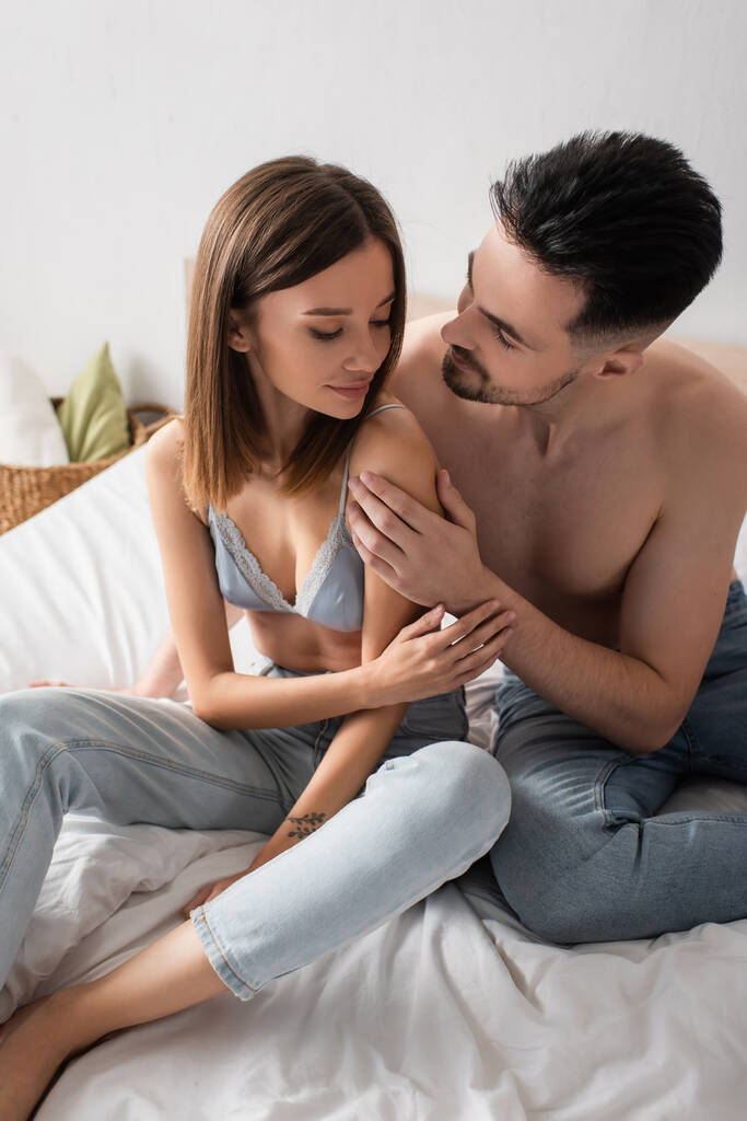 сексуальний молодий чоловік обіймає красиву жінку, сидячи на ліжку в джинсах і бюстгальтері
 - Фото, зображення
