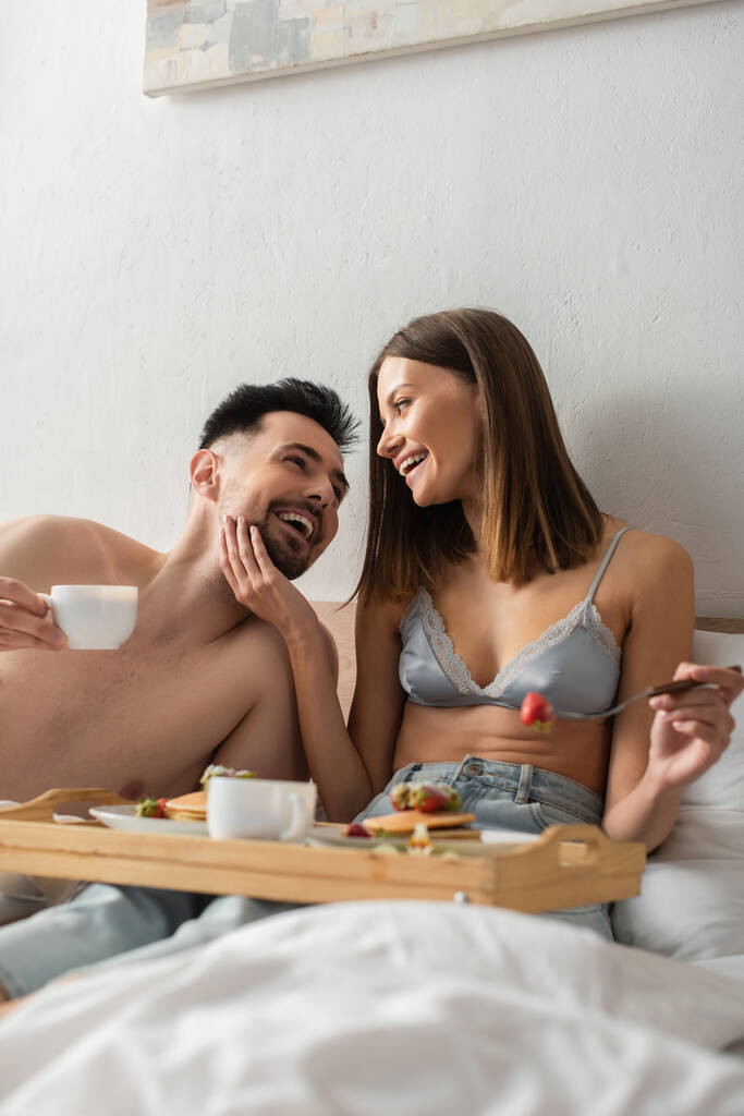 щаслива жінка в бюстгальтері, що торкається обличчя без сорочки хлопця під час сніданку в ліжку
 - Фото, зображення