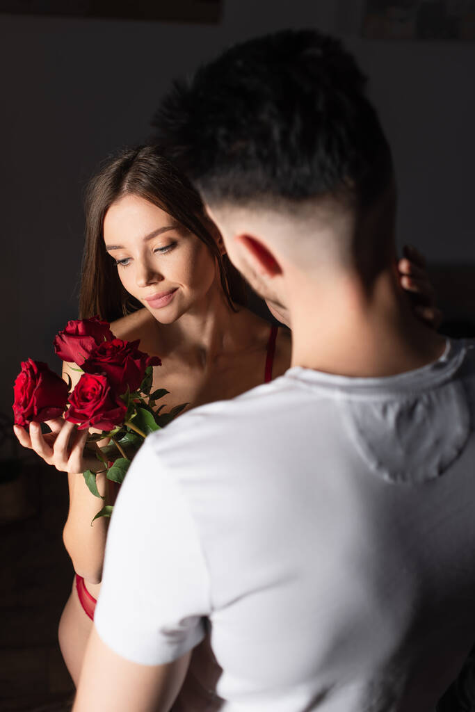 back view of blurred man in white t-shirt presenting red roses to sensual smiling woman - Foto, afbeelding