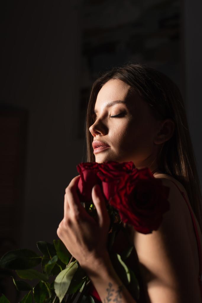 mujer joven y sensual con los ojos cerrados sosteniendo rosas rojas sobre fondo oscuro - Foto, Imagen