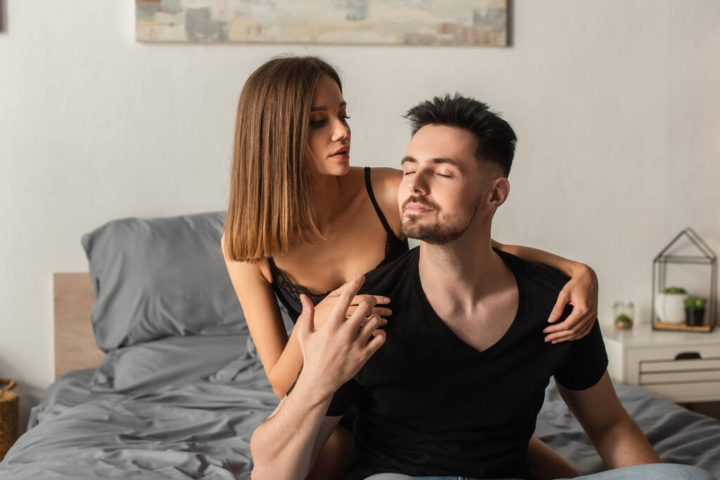 sensual young woman hugging man in black t-shirt sitting on bed with closed eyes - Fotoğraf, Görsel