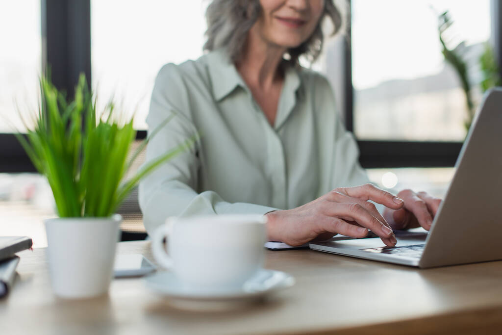 Gekropte weergave van wazig zakenvrouw met behulp van laptop in de buurt van koffie en plant in kantoor  - Foto, afbeelding