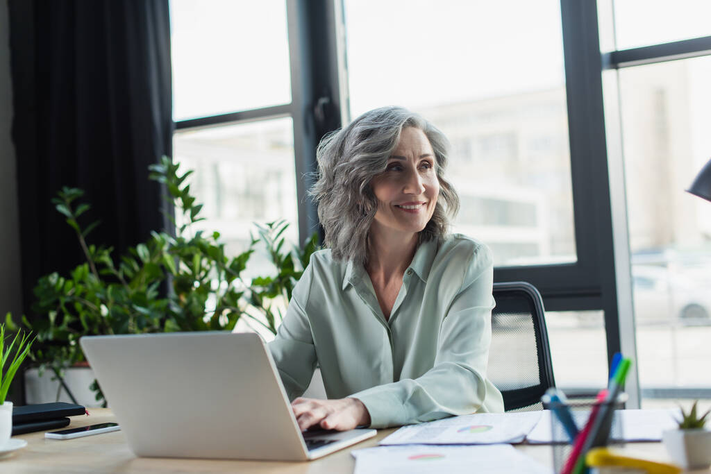 Усміхнена зріла бізнес-леді використовує ноутбук біля паперів в офісі
  - Фото, зображення