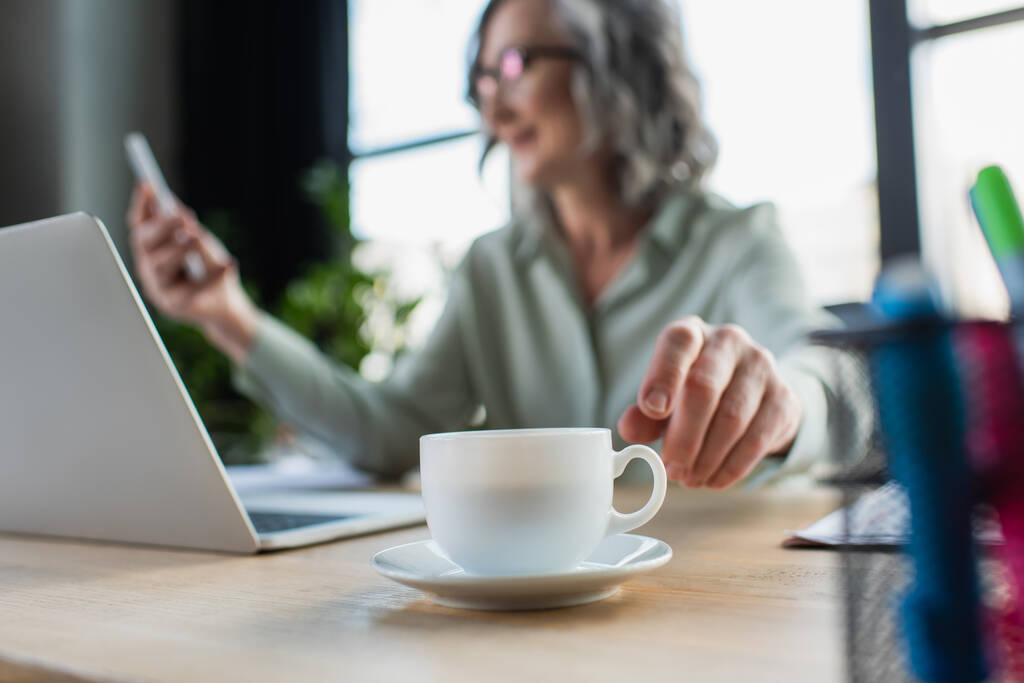 Чашка кави біля розмитої бізнес-леді і ноутбука в офісі
  - Фото, зображення