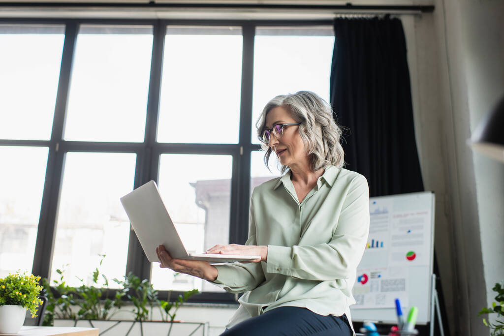 Зріла бізнес-леді в окулярах використовує ноутбук в офісі
  - Фото, зображення