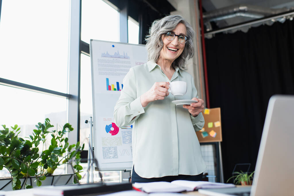 Mujer de negocios sonriente sosteniendo taza de café y mirando a la computadora portátil en la oficina  - Foto, imagen