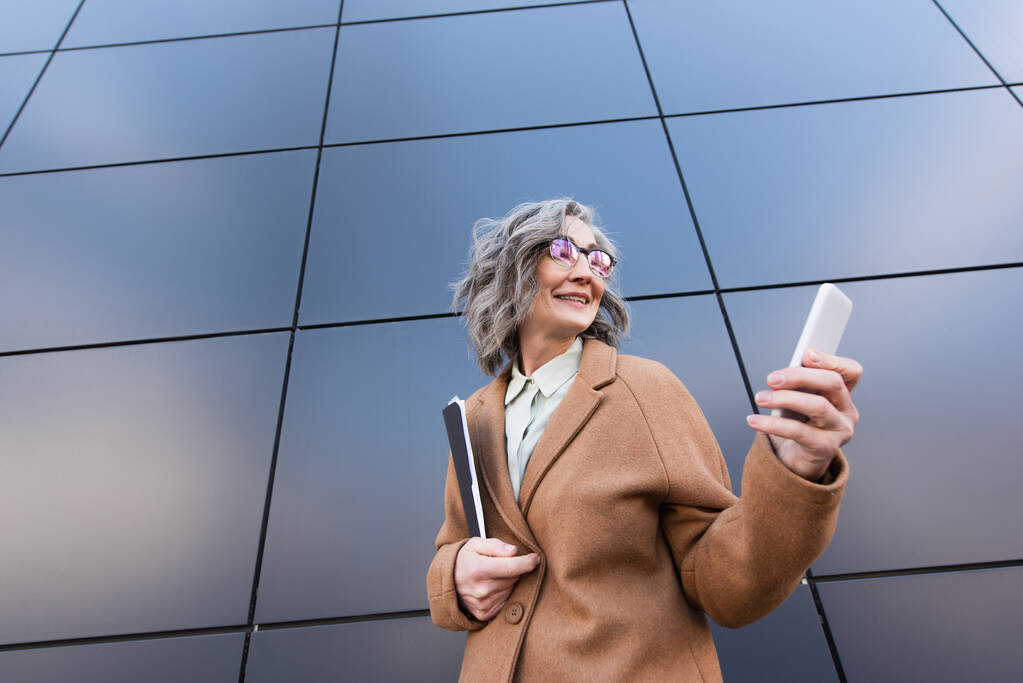 Низький кут зору весела зріла бізнес-леді в пальто, використовуючи мобільний телефон і тримаючи паперову теку біля будівництва на відкритому повітрі
  - Фото, зображення