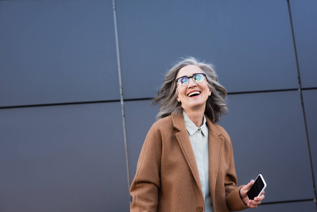 Femme d'affaires joyeuse en manteau et lunettes tenant smartphone avec écran blanc à l'extérieur  - Photo, image