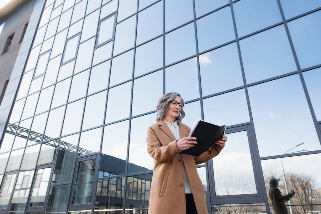 Vue en angle bas d'une femme d'affaires souriante en manteau regardant un dossier en papier et tenant un smartphone près d'un bâtiment à l'extérieur  - Photo, image