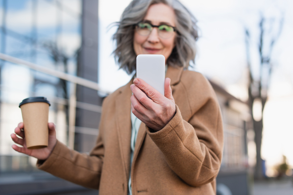 Smartphone in der Hand einer verschwommenen Geschäftsfrau im Mantel, die Kaffee hält, um ins Freie zu gehen  - Foto, Bild