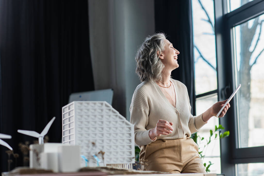Vista laterale di donna d'affari positiva che tiene il tablet digitale vicino a modelli di edifici in ufficio  - Foto, immagini