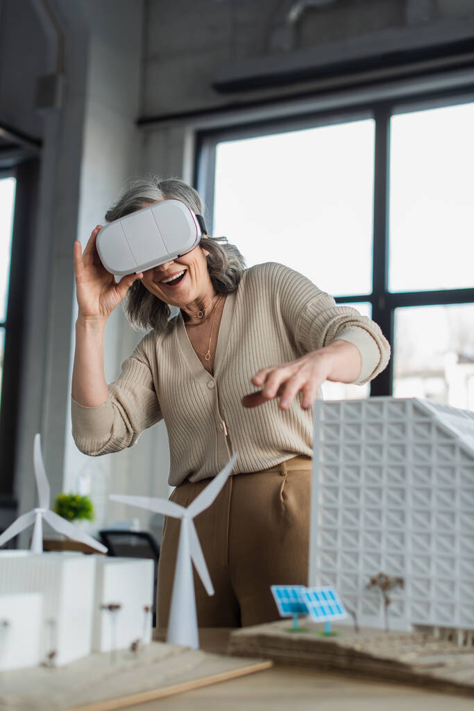 Счастливая деловая женщина в наушниках виртуальной реальности рядом с моделями зданий в офисе  - Фото, изображение