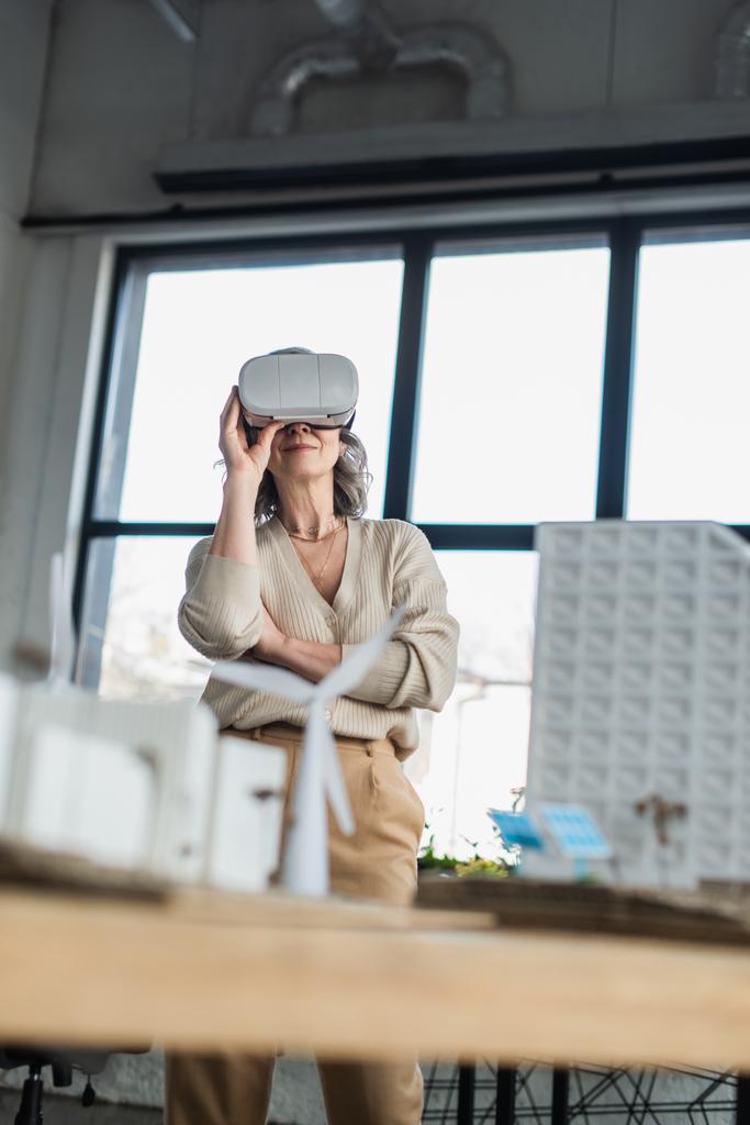 Femme d'affaires mature utilisant un casque vr près de modèles flous de bâtiments dans le bureau  - Photo, image