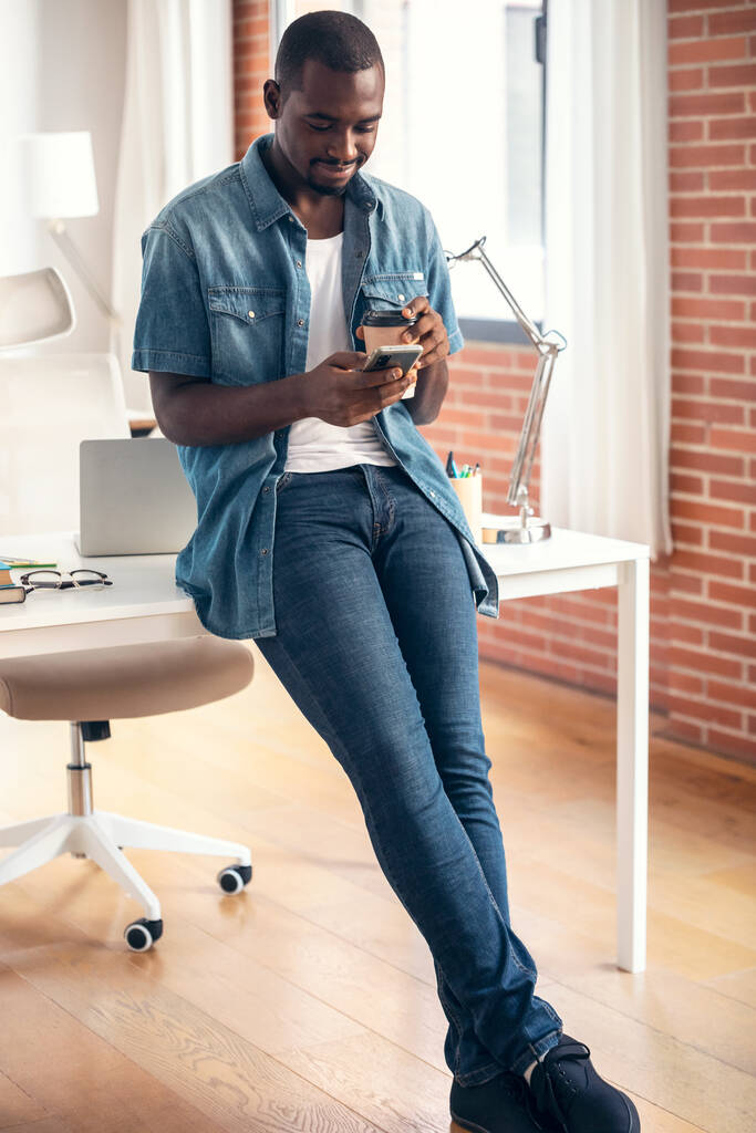Знімок щасливого бізнесмена, використовуючи смартфон, п'ючи чашку кави, сидячи на столі в коворкінгу
. - Фото, зображення