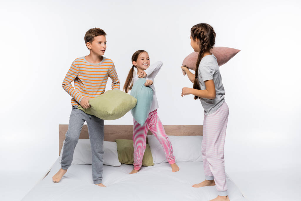 χαρούμενα παιδιά με πιτζάμες να μαλώνουν ενώ στέκονται στο κρεβάτι απομονωμένα στα λευκά - Φωτογραφία, εικόνα