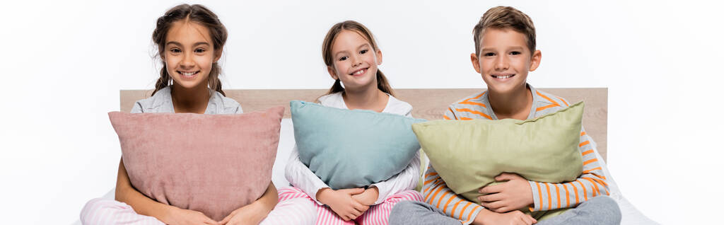 radostné děti sedící na posteli a objímající polštáře izolované na bílém, prapor - Fotografie, Obrázek