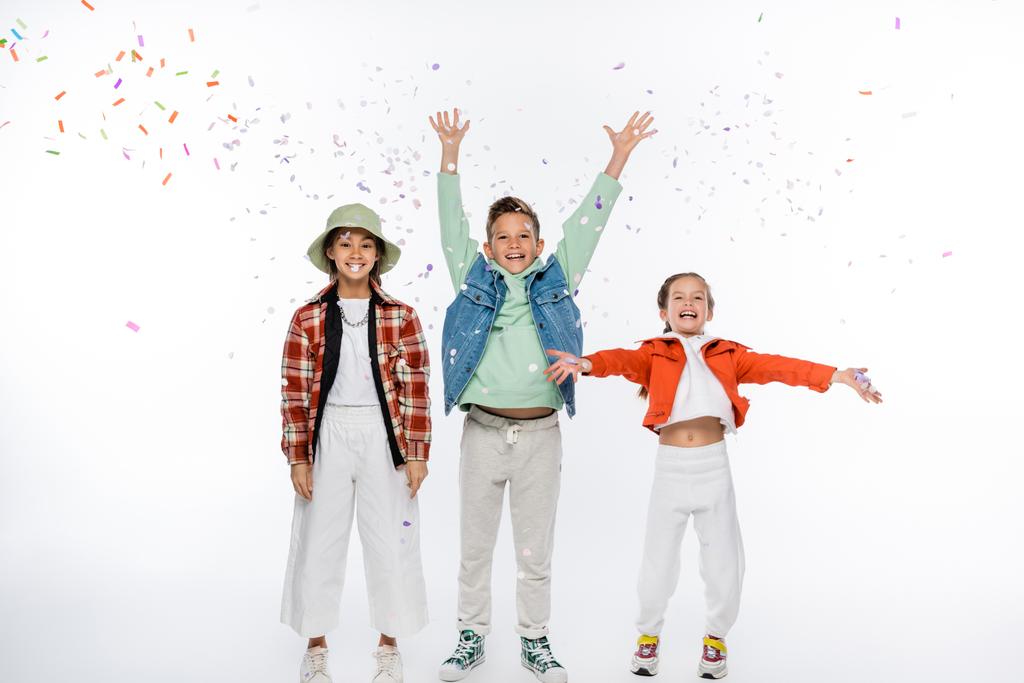полная длина положительного preteen мальчика с поднятыми руками, стоящих рядом с девушками и падения конфетти на белом - Фото, изображение