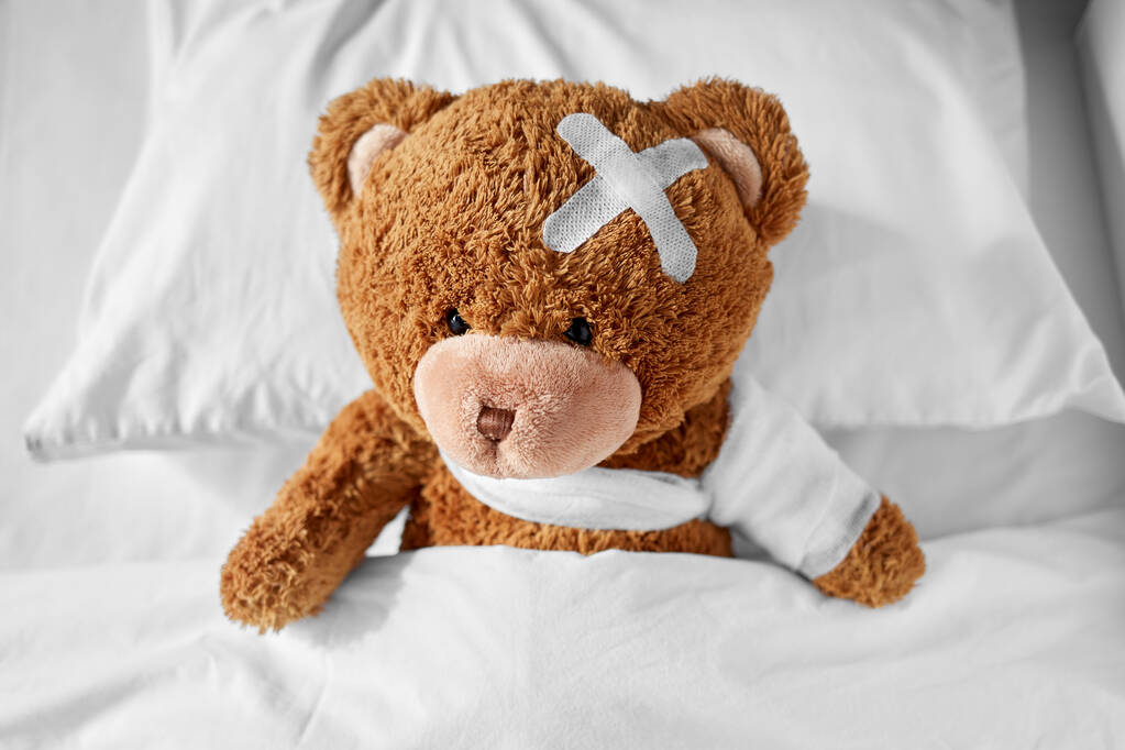 больной плюшевый мишка с перевязанной лапой и головой в постели - Фото, изображение