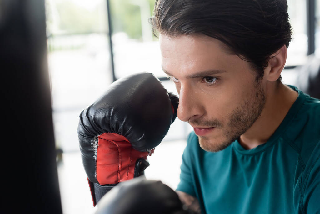Retrato de desportista em luvas de boxe olhando para saco de perfuração borrada no ginásio  - Foto, Imagem