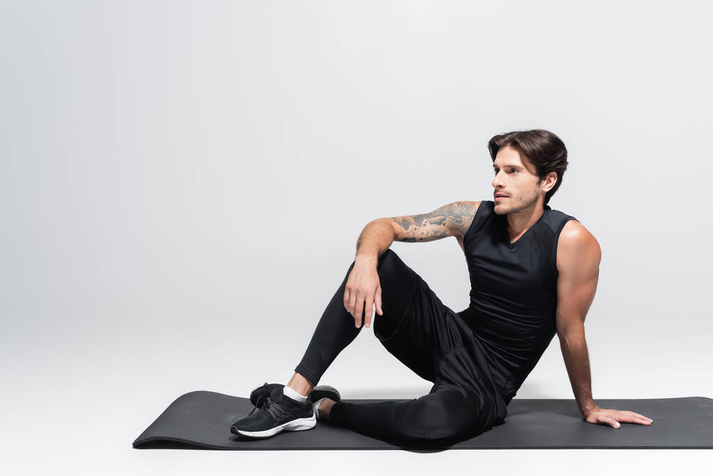 Мужчина сидит на черном фитнес-коврике на сером фоне - Фото, изображение