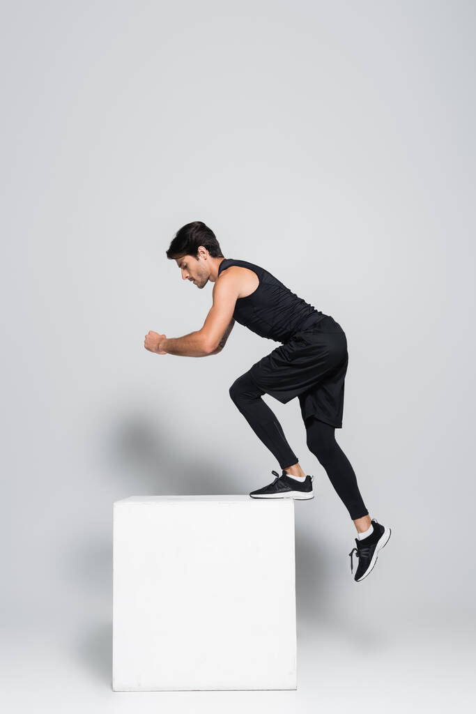 Vista lateral del deportista en ropa deportiva negra haciendo ejercicio con cubo sobre fondo gris  - Foto, imagen