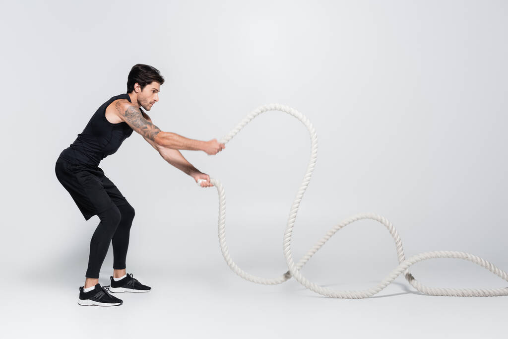 Side view of sportsman working out with battle ropes on grey background  - Photo, Image