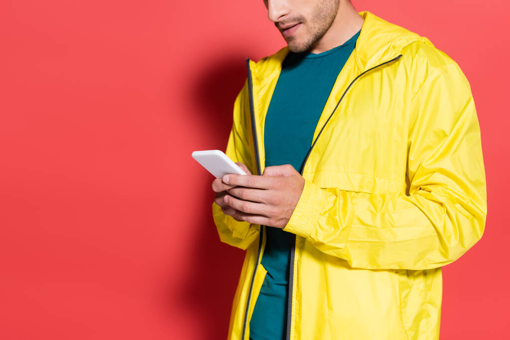 Cropped view of sportsman in yellow sports jacket using mobile phone on red background  - Photo, Image