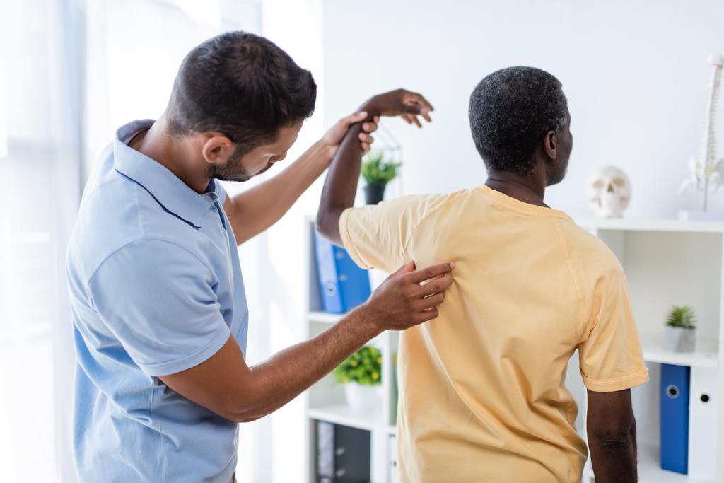 νεαρός θεραπευτής που εξετάζει την πλάτη ενός αφροαμερικανού ασθενή στο νοσοκομείο - Φωτογραφία, εικόνα