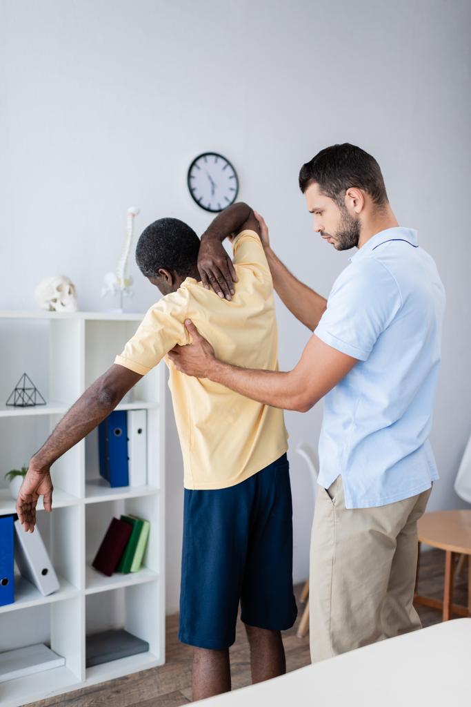 jonge arts aanraken van Afrikaanse Amerikaanse patiënt, terwijl de diagnose hem in revalidatiecentrum - Foto, afbeelding