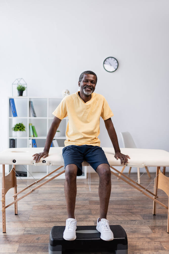 plný pohled na Afroameričana středního věku sedícího na masážním stole v rehabilitačním centru - Fotografie, Obrázek