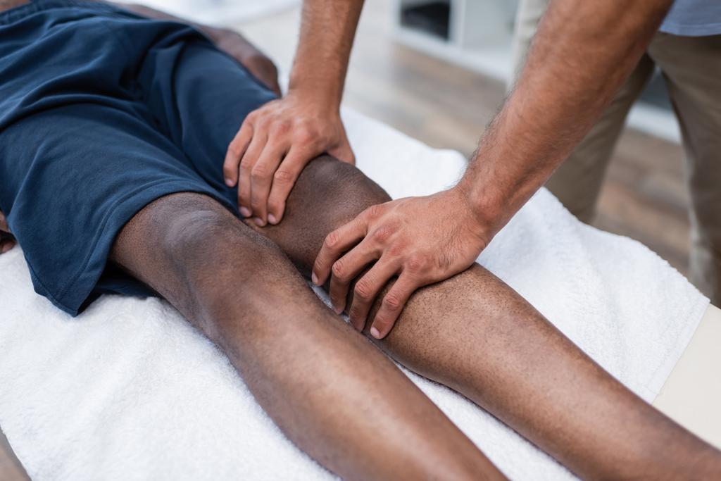 vista recortada de masajista masajeando rodilla de hombre afroamericano en centro de rehabilitación - Foto, Imagen