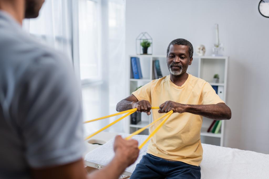 blurred rehabilitologist and african american patient doing exercise with elastics - Photo, Image
