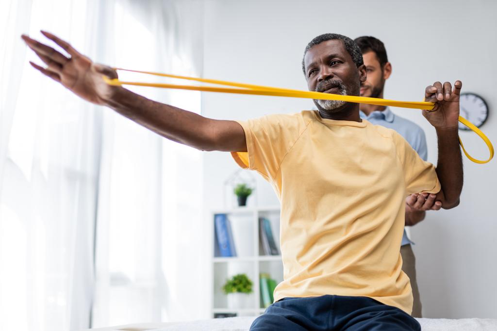 fysiotherapeut assisteren Afrikaans amerikaanse man uit te werken met elastiek - Foto, afbeelding