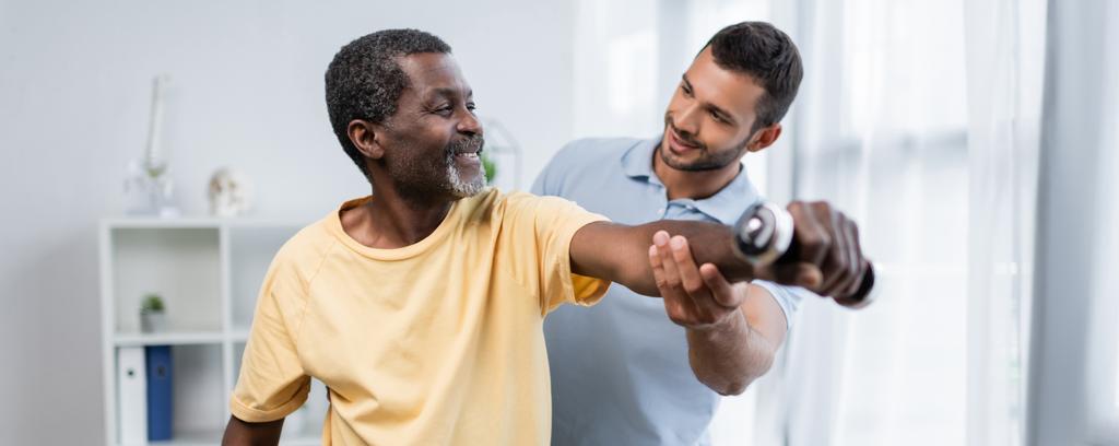 jovem fisioterapeuta assistindo sorridente paciente afro-americano exercitando com haltere, bandeira - Foto, Imagem