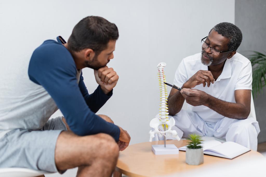 Африканский американский физиотерапевт указывает на модель позвоночника рядом с молодым человеком в консультационной - Фото, изображение