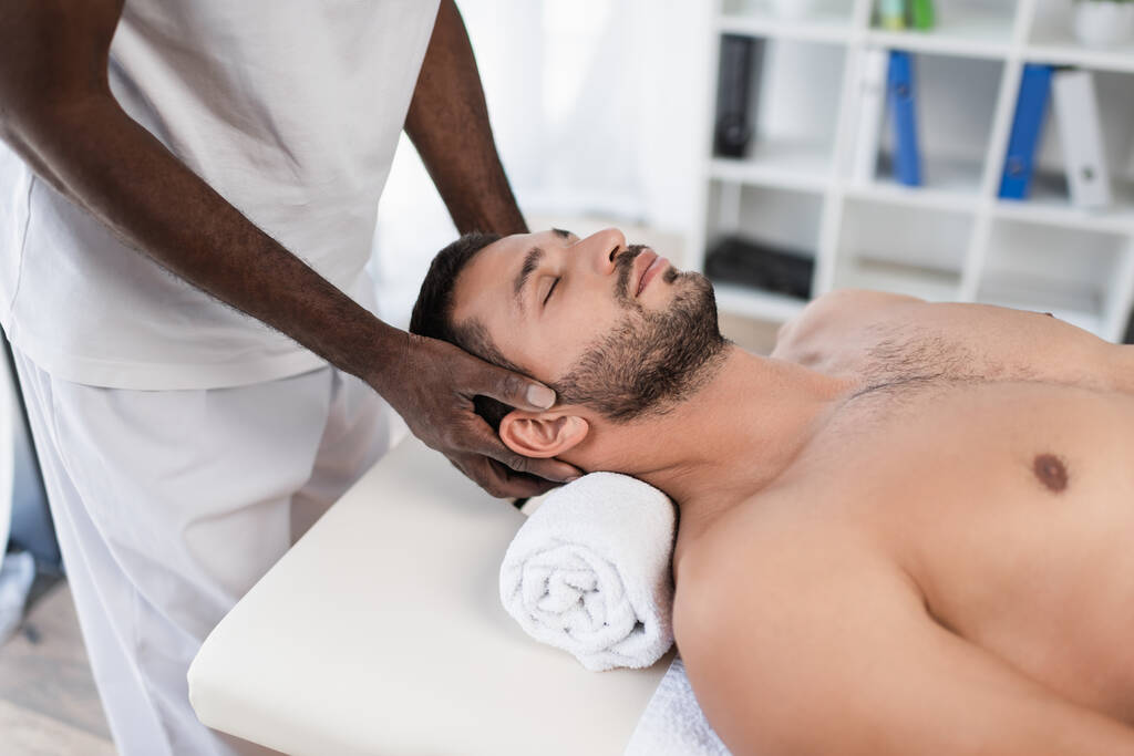 jonge man met gesloten ogen liggend op massage tafel tijdens revalidatie behandeling door afrikaanse amerikaanse masseur - Foto, afbeelding