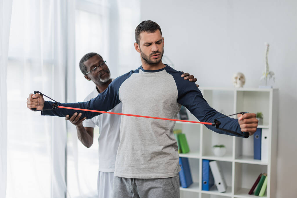 africký americký rehabilitolog pomáhá mladému muži trénovat s odbojovým pásmem  - Fotografie, Obrázek