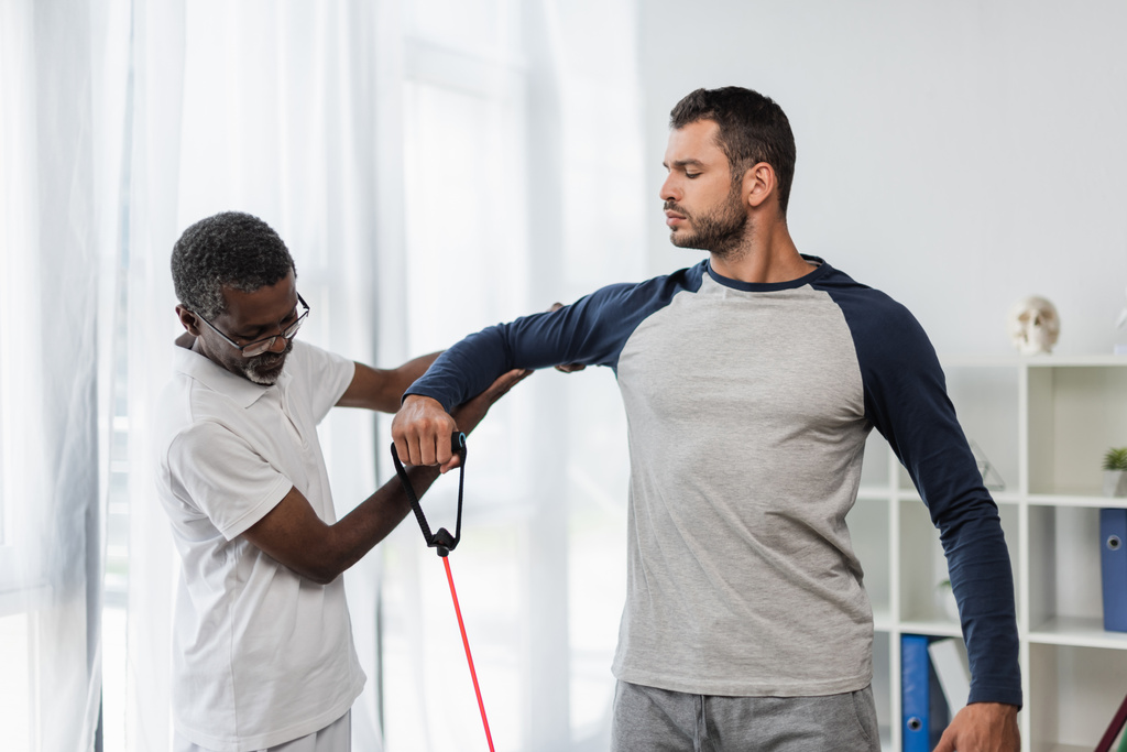 zralý africký americký trenér pomáhá mladému muži cvičit s odbojovým pásmem v rehabilitačním centru - Fotografie, Obrázek