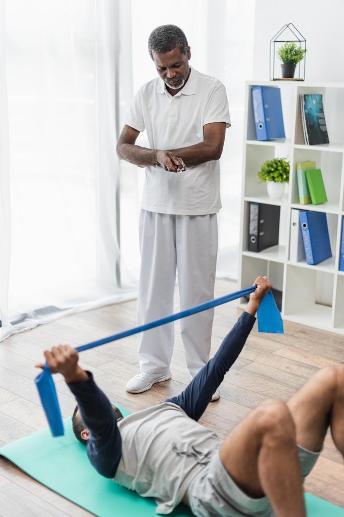 Afrikaans amerikaanse rehabilitoloog op zoek naar jonge man training met rubber band terwijl liggend op fitness mat - Foto, afbeelding