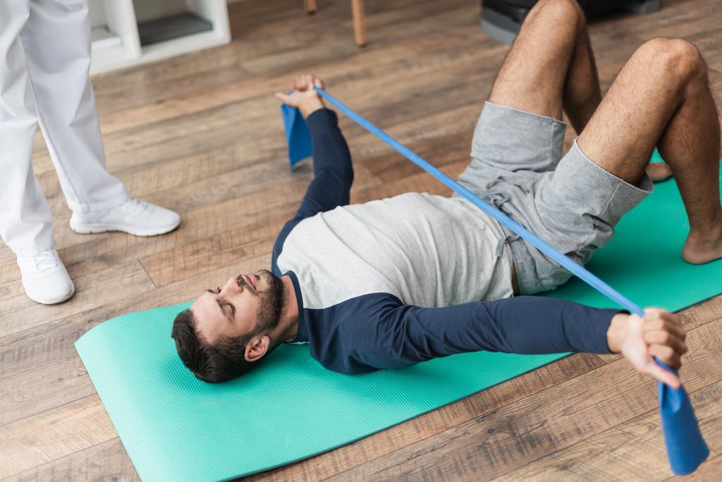 fiatal férfi fekvő fitness mat és képzés gumiszalaggal közel rehabilitológus - Fotó, kép